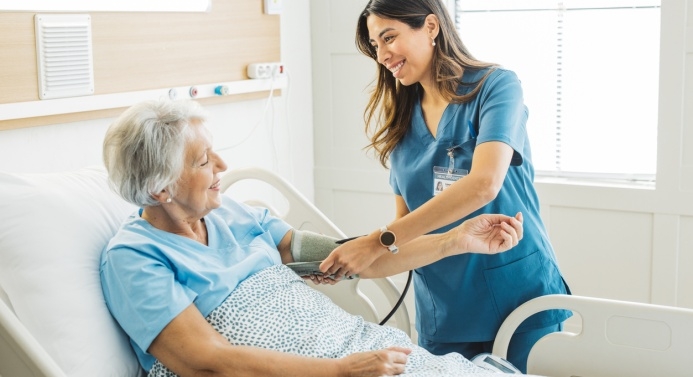 Hoitaja mittaa verenpainetta ikääntyneeltä sairaalavuoteessa makaavalta nainen.