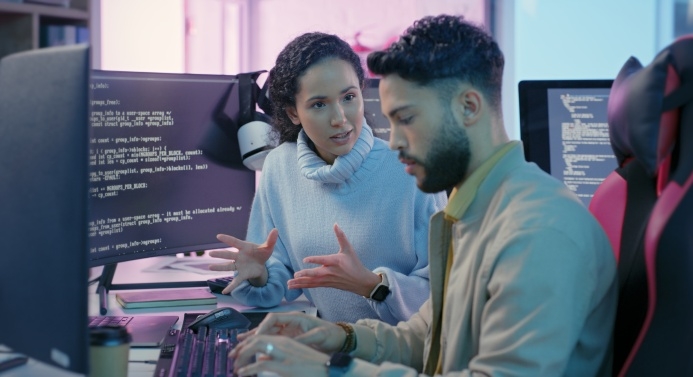 Nainen neuvoo tietokoneella työskentelevää miestä, taustalla tietokoneruutuja.